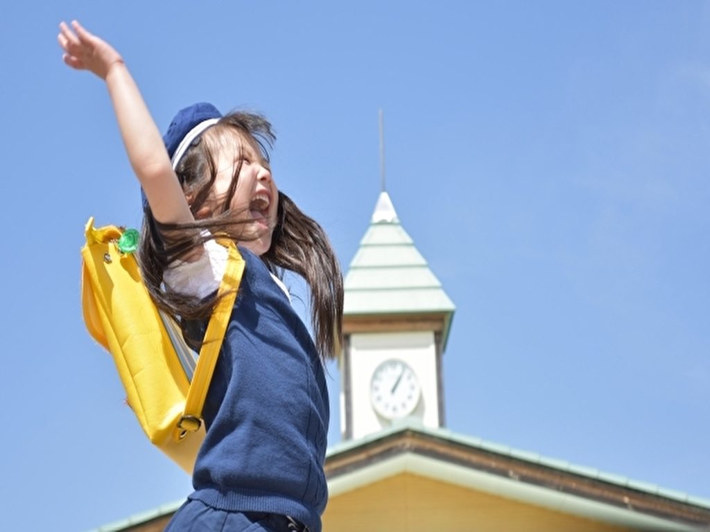 青空のもと校舎の前で黄色いリュックを背負って制服を着た女の子の園児がジャンプしているところ